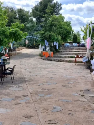 Eventos el regajo Vista general del patio exterior de finca El Regajo decorado para celebración, con terraza de losas de piedra, mesas y sillas bajo los árboles, escalinata central con decoración de telas blancas y tinaja de barro en primer plano junto a fachada de piedra.